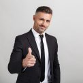 Portrait of a handsome confident businessman wearing suit standing isolated over gray backgorund, showing thumbs up
